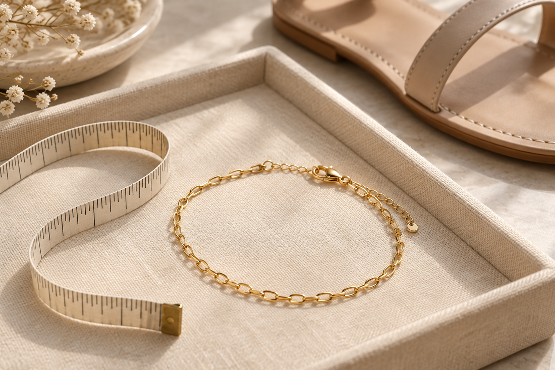 Yellow-gold link anklet with a clasp and extender on an ivory tray beside a measuring tape and beige sandal.