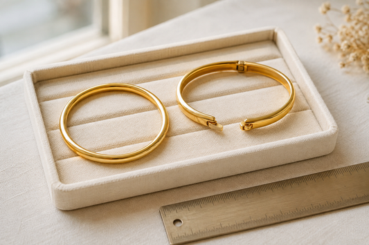 Round slip-on gold bangle and open hinged gold bangle on an ivory tray beside a ruler.