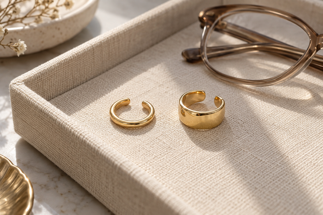 Two yellow-gold ear cuffs with open gaps on an ivory linen tray beside folded eyeglasses.