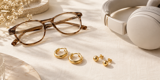 Gold hoop and stud earrings on ivory linen beside folded glasses and over-ear headphones.