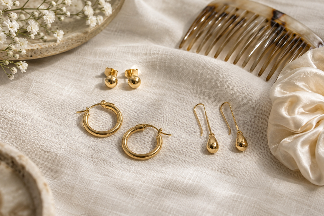 Gold stud, hoop and drop earrings on ivory linen beside a hair comb and satin hair tie.