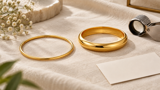 Two plain yellow-gold bangles of different thicknesses on ivory linen beside a jeweller’s loupe and blank card; illustrative image.