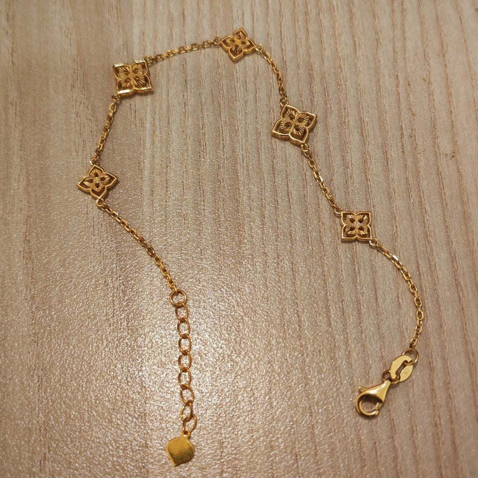 A gold chain bracelet with five small square quatrefoil motifs and an adjustable extender chain with a lobster clasp, displayed on a light wood grain surface.
